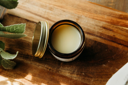 Open jar of Bohemian Rebloom Herbal Tallow Balm on a wooden surface with green leaves.